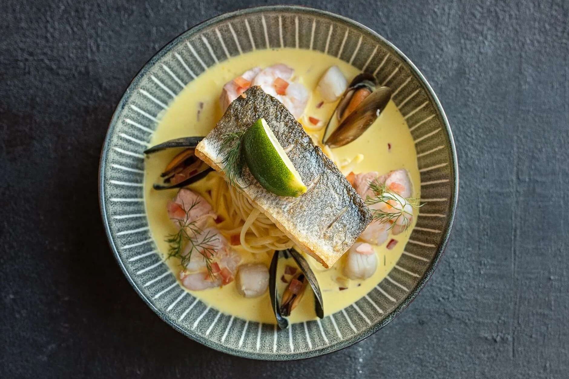 overhead shot of the Glenesk Hotel food from Fairways Brasserie and Grill Fish Dish with mussels, sauce and lime wedge on top set against a grey background.