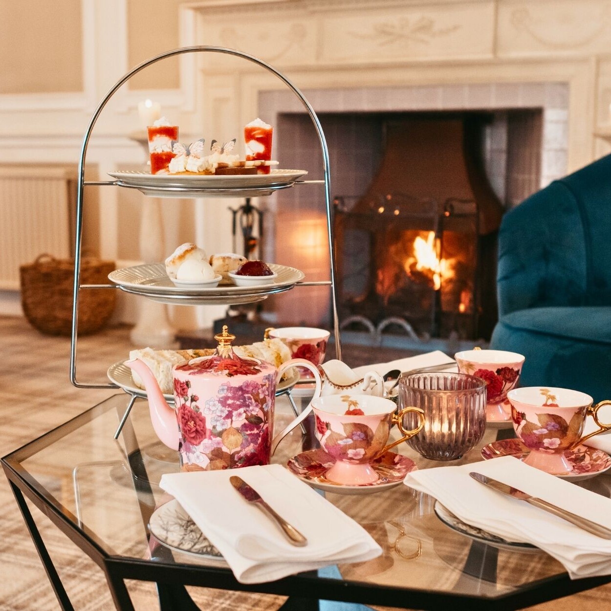 Coffee table with afternoon tea and beautiful pink china teacups and teapot. place setting with knoives and napkins. Blue sofas, burning log fire and fireplace in background.