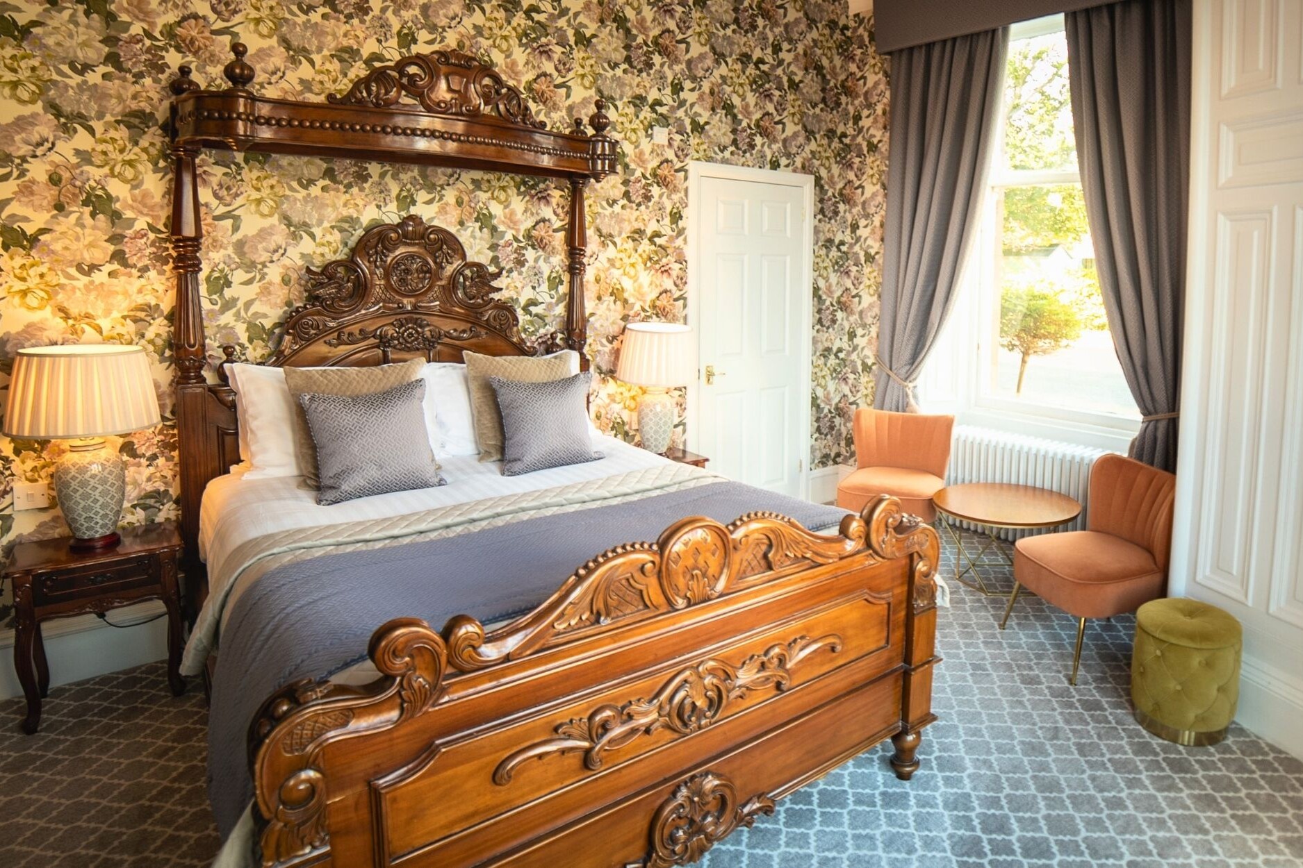 Beautfiful ornate carved wooded bed and decorative cushions and side lamps. Orange velvet chairs and coffee table. Luxury floral wallpaper and grey curtains with tie backs.