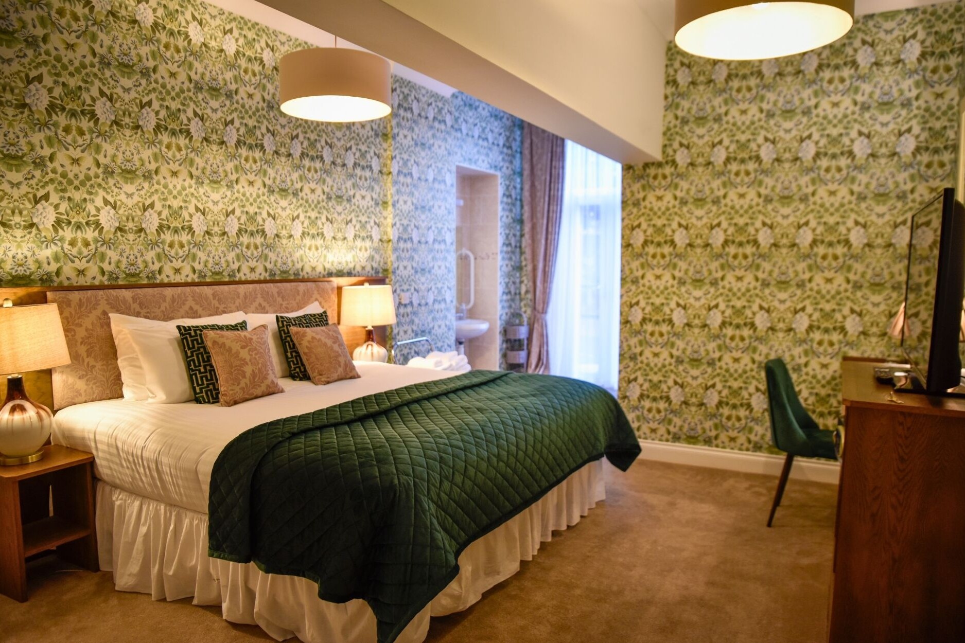 Hotel room shot of the accessible room at the Glenesk Hotel, Large king size bed, tv and dressing table chair. sink area. Decorative floral wallpaper.