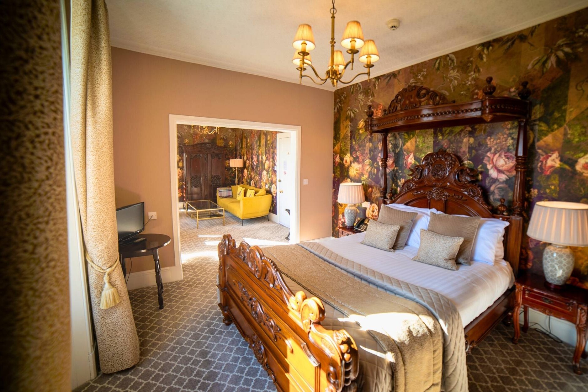 Beautiful luxury ornate wooden bed with decorative cushions, floral wallpaper and side lamps looking through to the seating area of suite with yellow sofa, coffee table and large ornate wardrobe