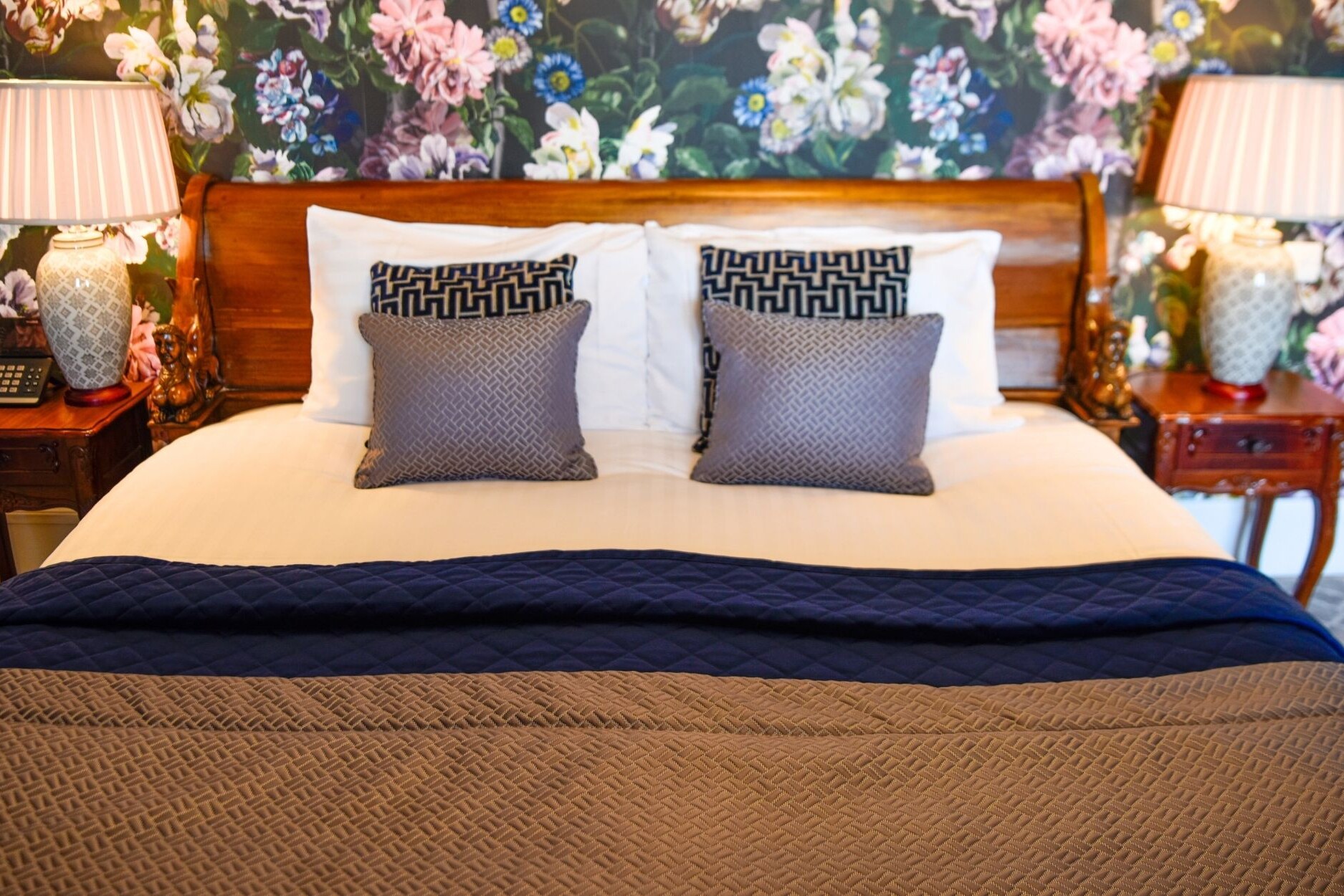 Junior Suite Bed Details. Wooden sleigh bed headboard with decorative cushions, luxury floral wallpaper and