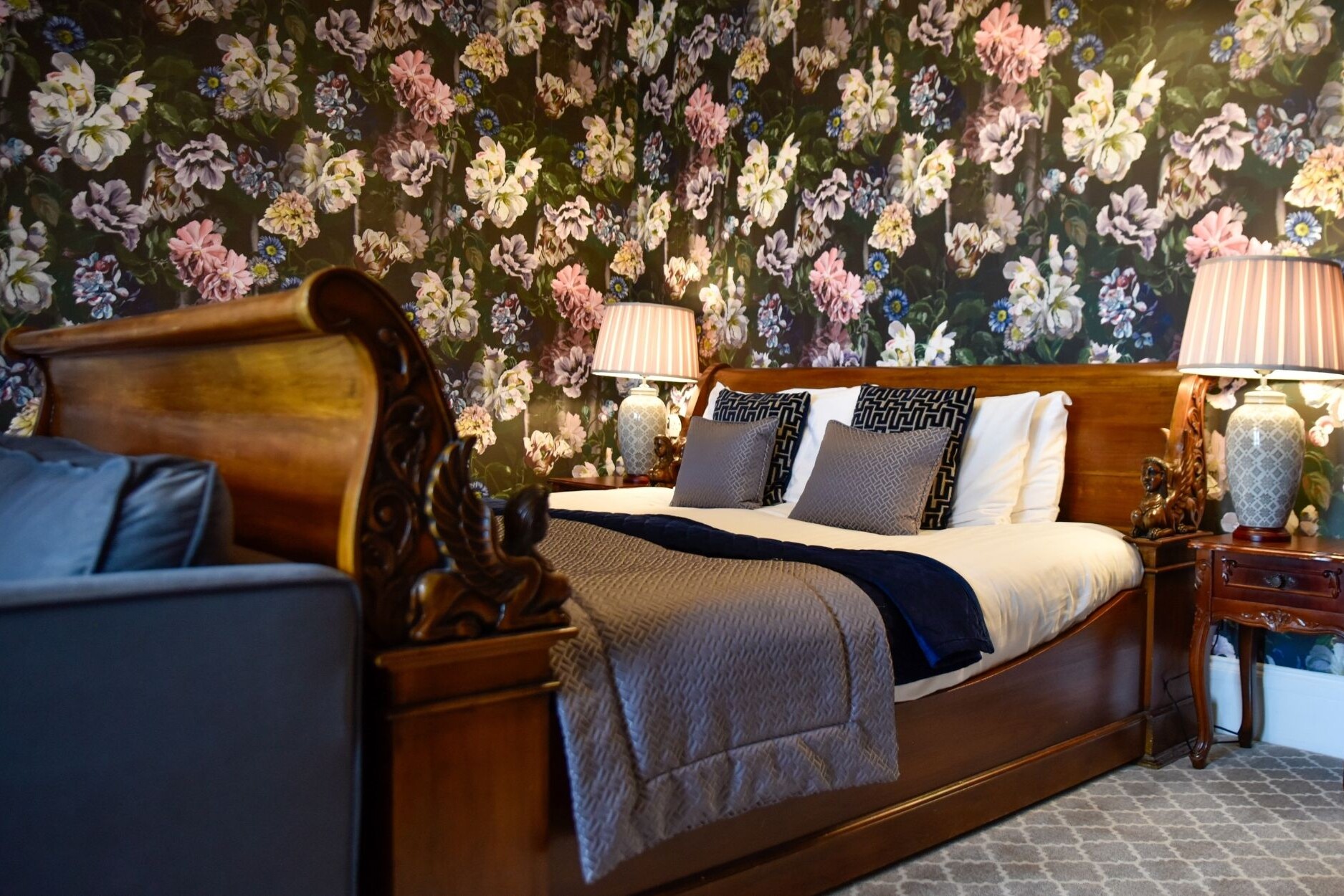 Side angle of the luxury wooden sleigh bed with ornate details in Junior Suite at the Glenesk Hotel. Luxury floral wallpaper, and feature side lamps