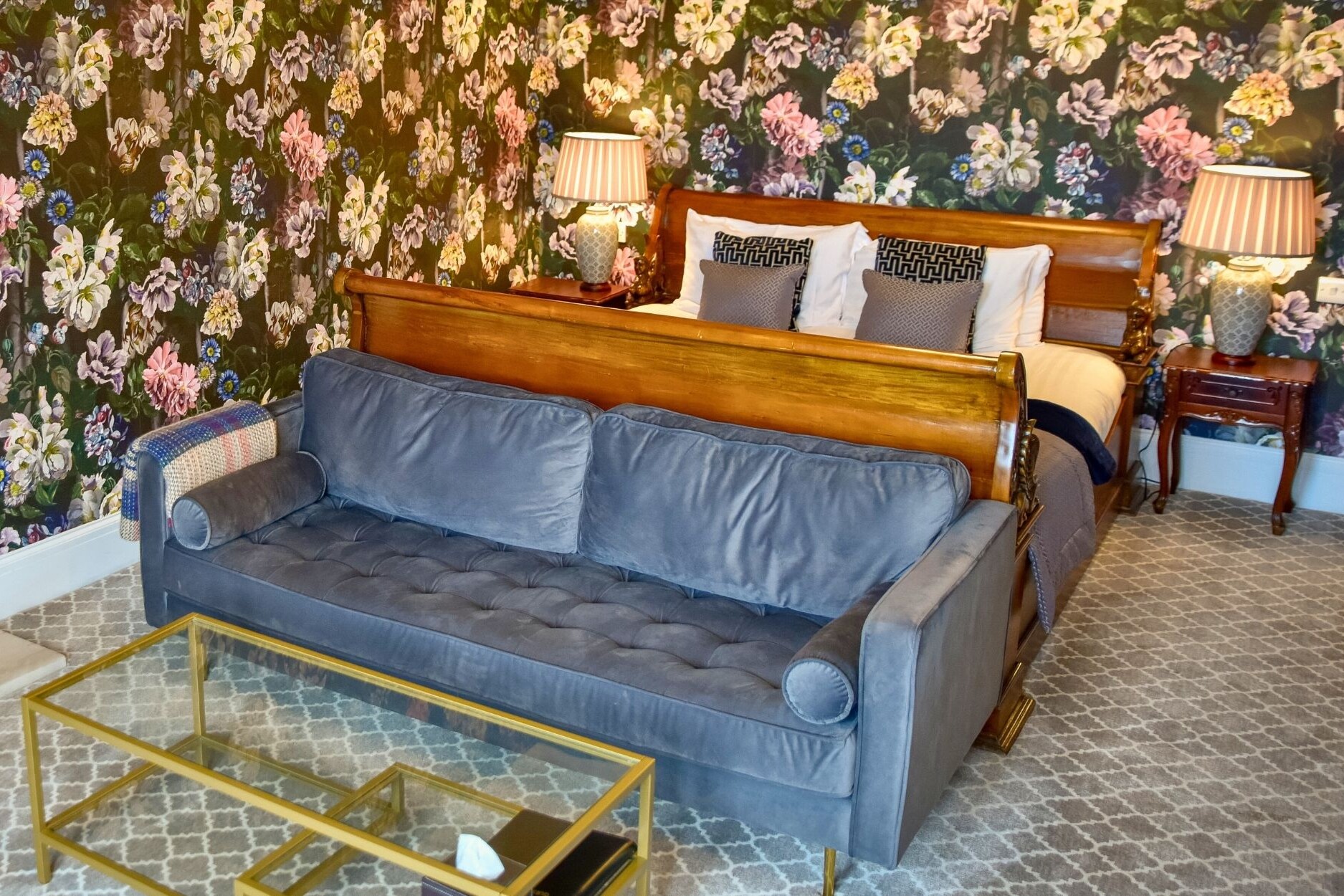 View from bottom of sleigh bed. Sofa and coffee table at bottom of bed, two side lamps and luxury floral wallpaper.