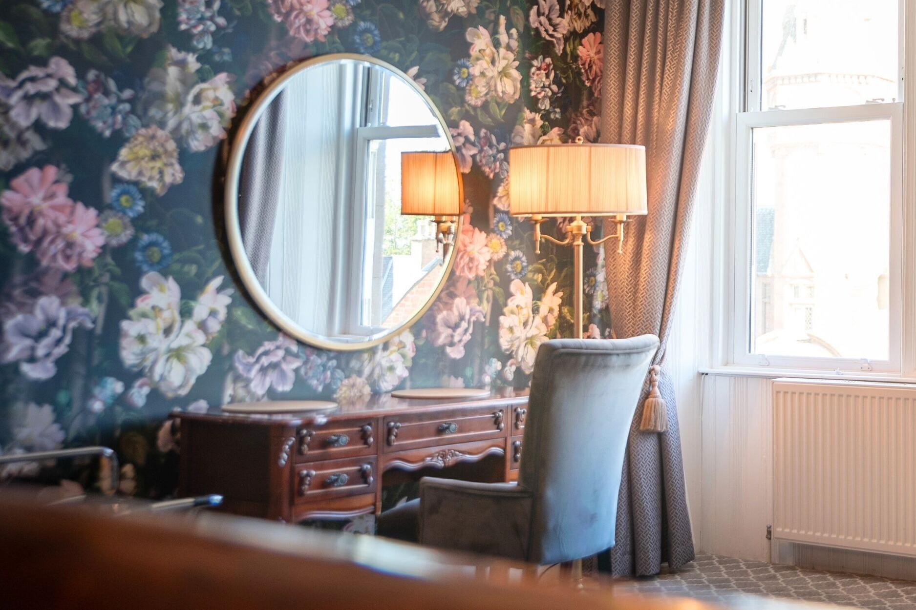 Dressing table, chair, round mirror and floor lamp with luxury floral wallpaper on walls.