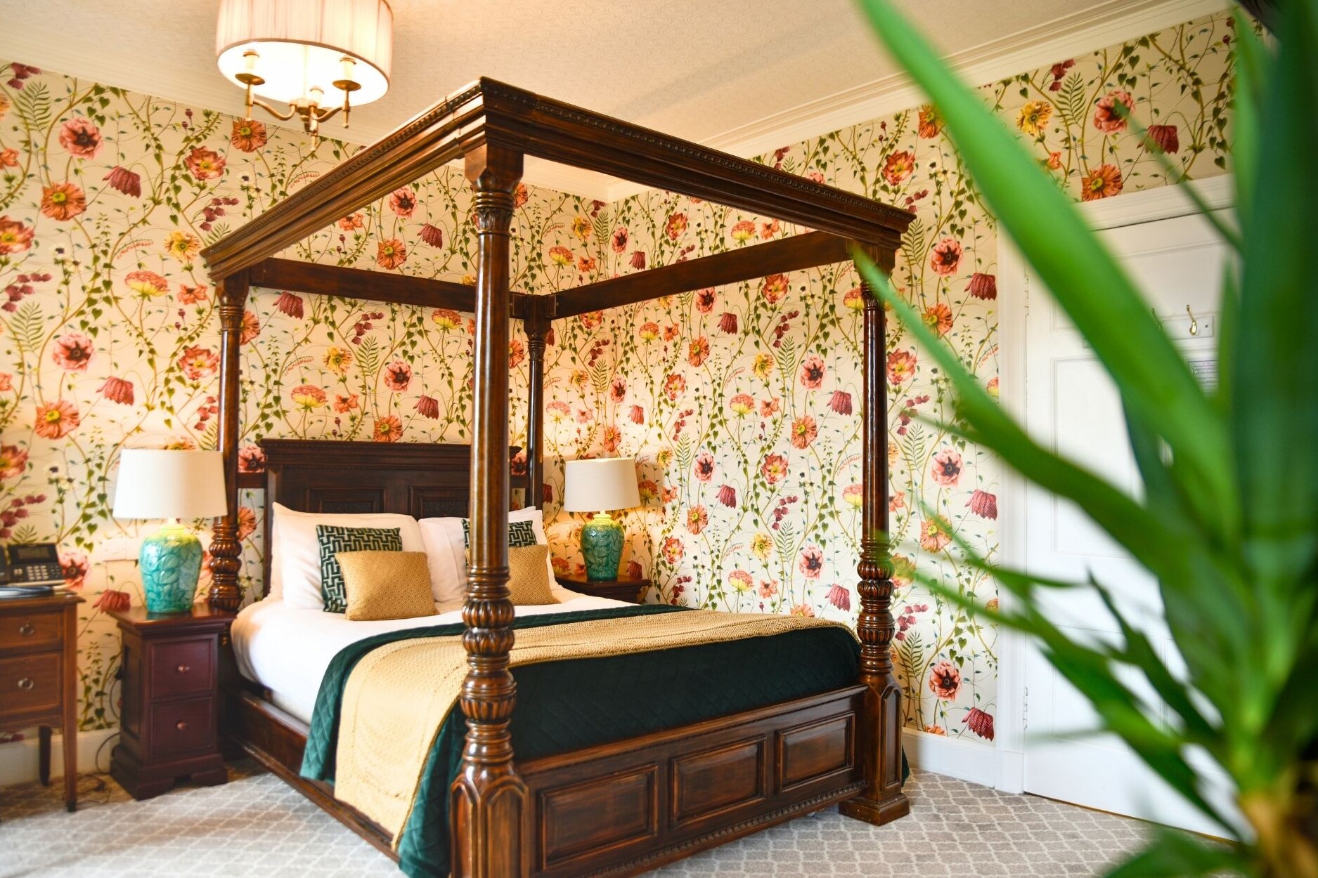 Beautiful wooden four poster bed with decorative cushions and side lamps. Luxury floral wallpaper.