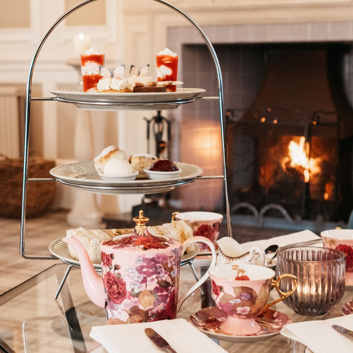 Afternoon tea stand with cakes and sandwiches on coffee tbalel with china teapot and cups sitting in front of the warm burning fireplace