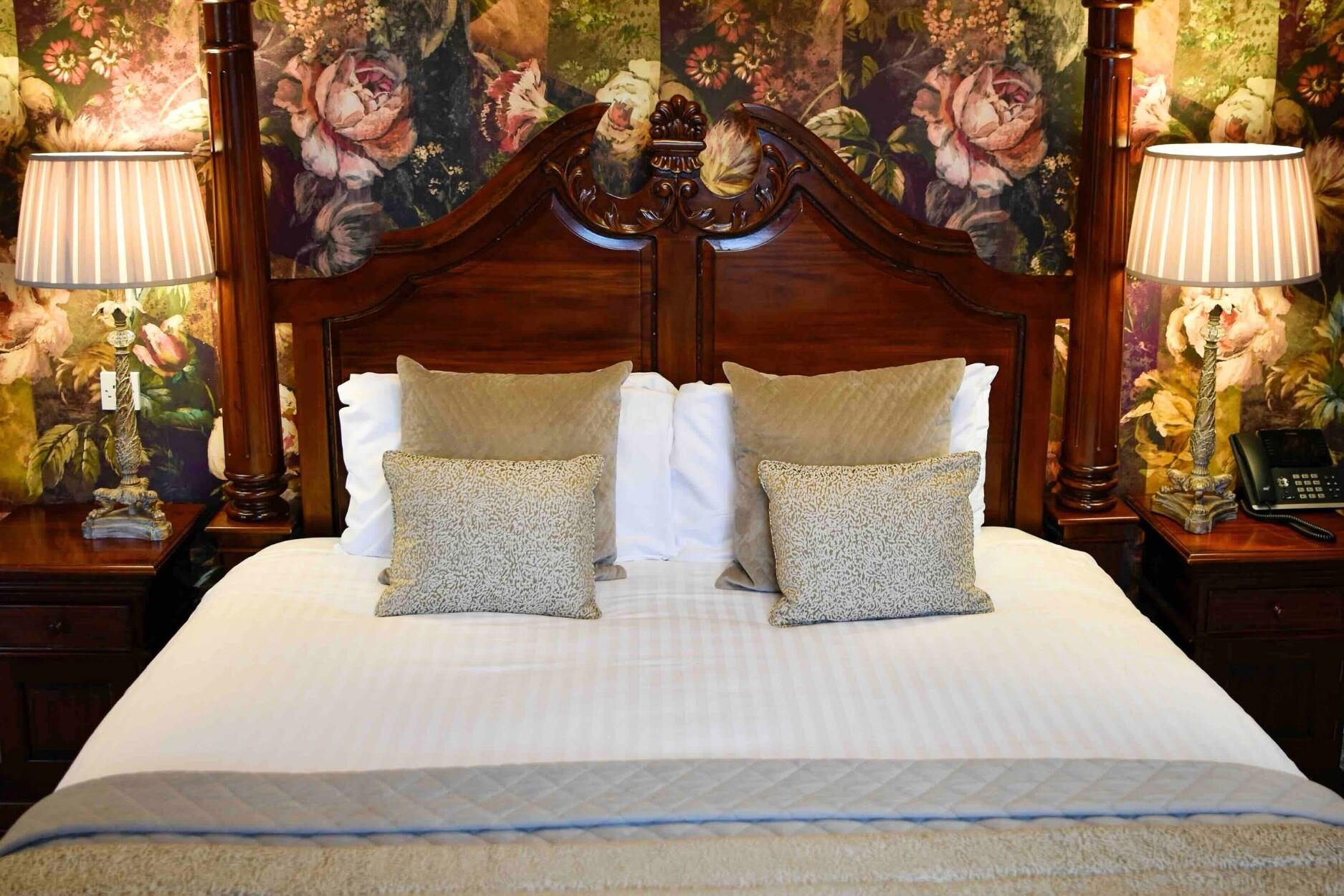 Four poster headboard close up with decorative cushions and side lamps against floral wallpaper